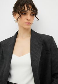 Blazer nero con righe sottili e rever a punta, abbinato a un top bianco senza maniche. Il blazer presenta spalline e una vestibilità classica.