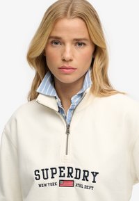 Blondynka o niebieskich oczach, ubrana w białą bluzę zapinaną na suwak z napisem "Superdry New York ATHL DEPT" i emblematem amerykańskiej flagi.