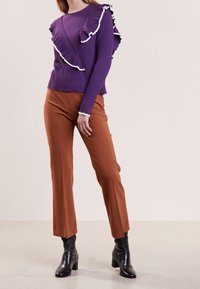 Haut violet à manches longues côtelées avec des accents blancs volantés, associé à un pantalon marron à jambes larges et des bottines noires à talons.