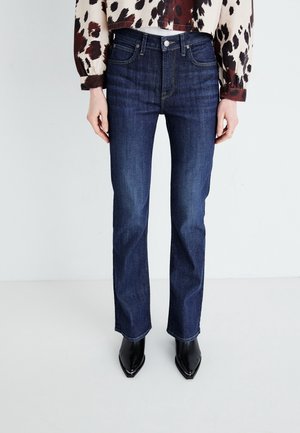 Vrouw draagt donkerblauwe jeans met rechte pijpen, een blouse met dierenprint en zwarte laarzen met puntige neus, staat tegen een effen lichte achtergrond.