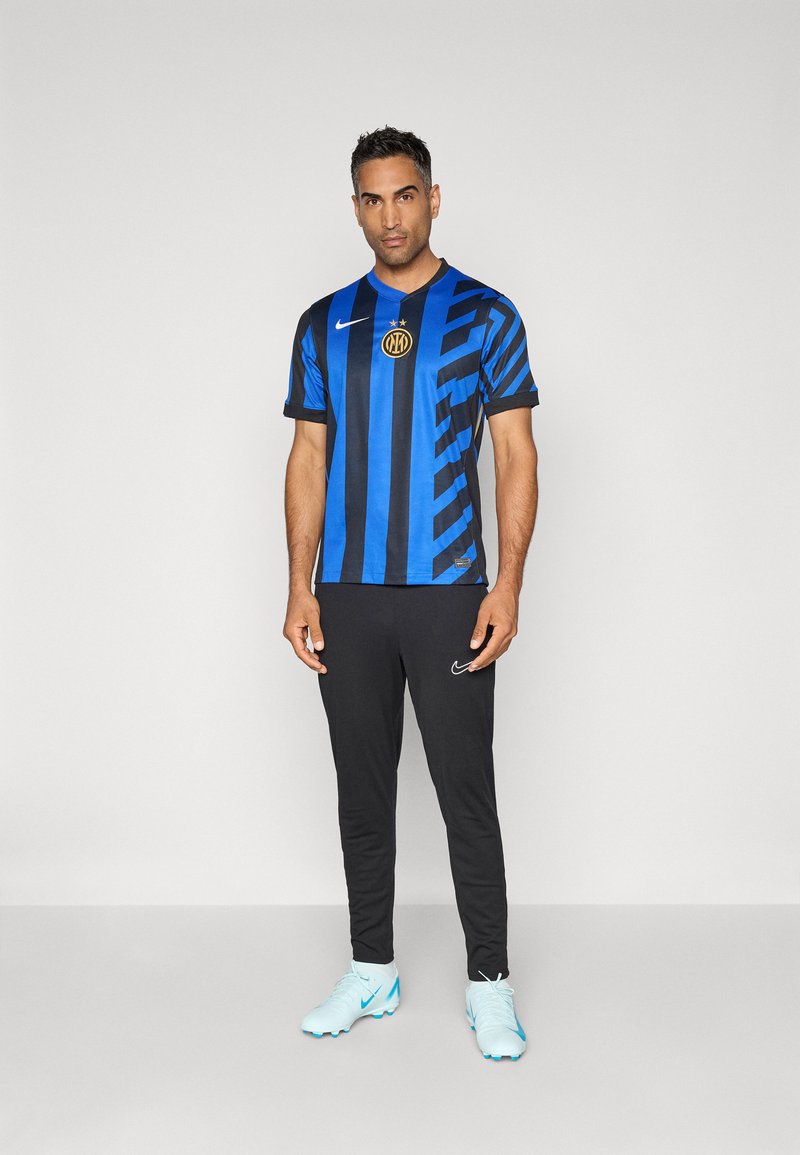 Hombre vistiendo una camiseta de fútbol a rayas azul y negra, pantalones negros y zapatillas de deporte celestes, de pie contra un fondo gris liso.