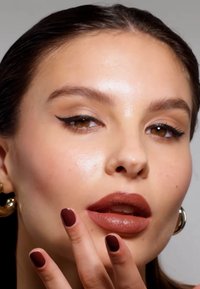 Närbild av ett ansikte med långt brunt hår, matta bruna läppar, mörk vinge-eyeliner och mörkröda naglar. Guldhoopörhängen är synliga.