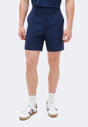 Homme portant un short bleu marine, des chaussettes blanches montantes et des baskets blanches avec des bandes rouges et bleues, debout devant un fond uni.