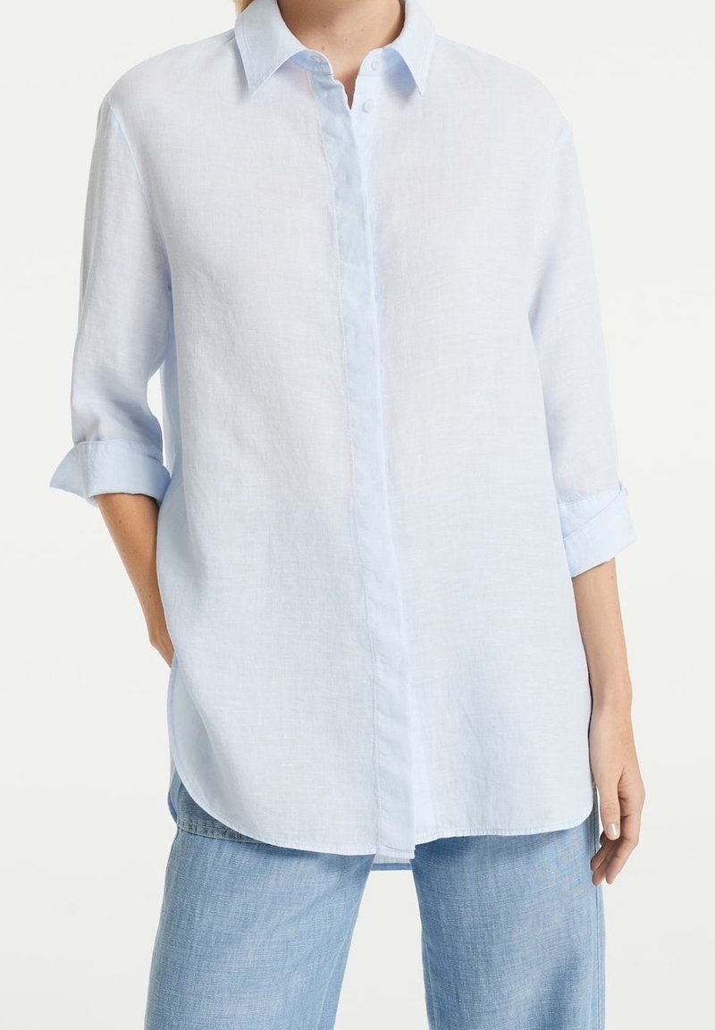 Personne portant une chemise bleu clair à manches longues avec les poignets retroussés et un jean bleu, debout avec une main dans la poche sur un fond blanc.