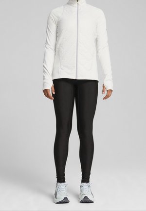 Person iført en hvid quiltet lynlåstrøje, sorte leggings og hvide løbesko, stående mod en ensfarvet grå baggrund.