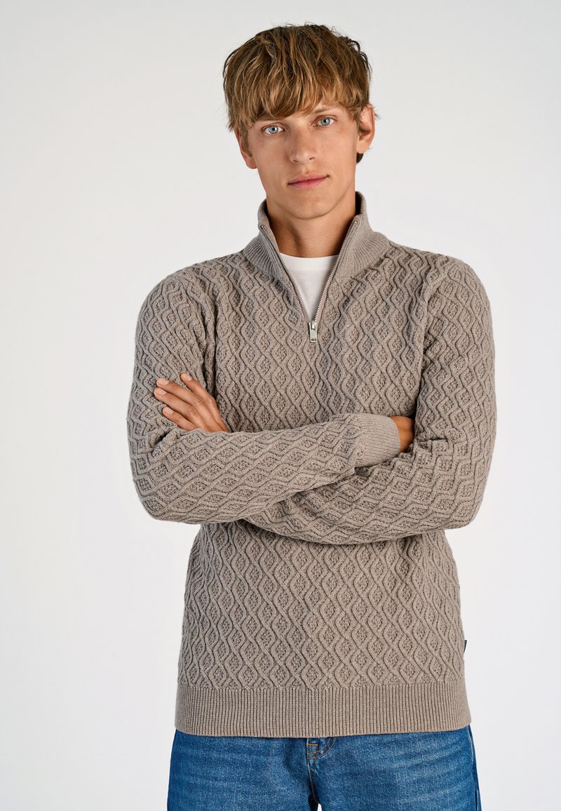 Lichtbruin gebreide trui met een ruitpatroon, met ritssluiting bij de kraag en geribbelde manchetten. Draag het met een witte t-shirt en blauwe jeans.
