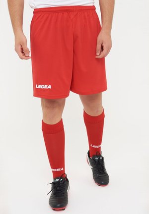 Personne portant un short de sport rouge, des chaussettes rouges montantes assorties et des crampons noirs avec des accents blancs, debout sur un fond blanc.