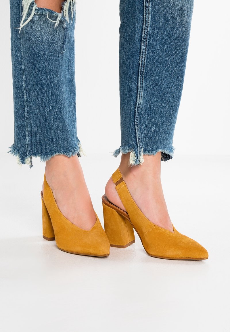 Pieds portant des escarpins à talons carrés en daim jaune moutarde avec brides arrière, associés à un jean en denim bleu à ourlet effiloché sur fond blanc.