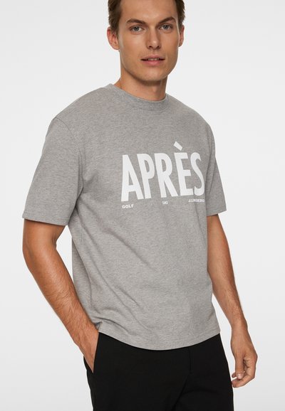 Mężczyzna ubrany w szary, krótki rękaw T-shirt z białym napisem "APRÈS" oraz mniejszymi słowami "GOLF" i "SKI", stojący z jedną ręką w kieszeni.