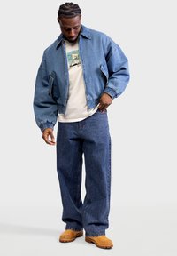 Giacca di jeans blu con polsini elastici, abbinata a una t-shirt bianca con stampa grafica e jeans blu larghi. L'outfit è completato da stivali marroni.