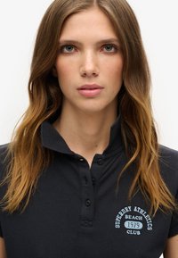 Camisa polo negra con cuello clásico, tres botones y texto bordado en azul: "Superdry Athletics Beach Club 1979."