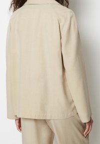 Beżowa, lekka kurtka o luźnym kroju, prostej linii dołu i raglanowych rękawach. Gładka faktura z minimalistycznym wzornictwem, bez widocznych elementów metalowych.