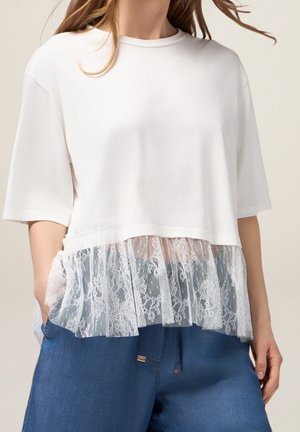 White short-sleeve top featuring a lace trim at the hem. The lace is soft and floral-patterned, layered over a light fabric.