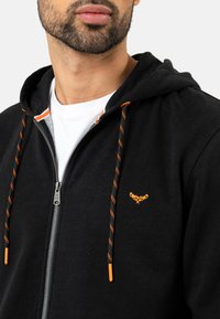 Sort zip-up hoodie med orange snor og et lille broderet design på brystet. Lavet af blødt materiale med en glat tekstur.