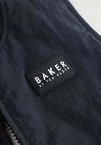 Juoda audinio medžiaga su siūta juoda stačiakampė lopinėliu, ant kurio baltomis didžiosiomis raidėmis užrašyta „BAKER BY TED BAKER“.