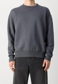 Sweatshirt gris en matériau doux avec un col rond, des poignets et un ourlet côtelés, arborant un petit logo sur la poitrine.