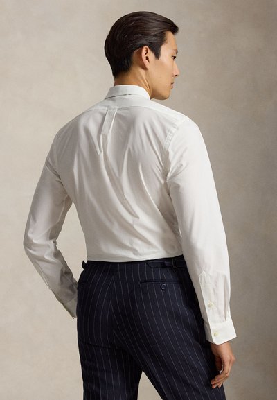 Man die een aansluitend wit overhemd en donkere pantalon met fijne strepen draagt, met zijn rug naar de kijker gekeerd en zijn hoofd in profiel tegen een neutrale achtergrond.