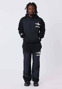 Conjunto de sudadera con capucha y pantalones negros con patrones irregulares de color claro. La tela parece suave y tiene un ajuste relajado. El modelo está de pie con las manos en los bolsillos.