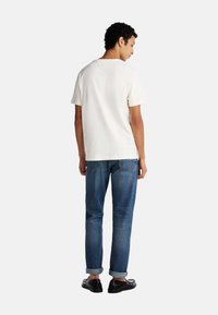 T-shirt bianco in cotone con maniche corte, abbinato a jeans in denim blu. I jeans presentano orli arrotolati e un dettaglio sulla tasca posteriore. Scarpe nere completano il look.