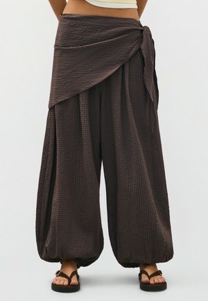 Personne portant un pantalon enveloppant marron texturé, ample, avec des poignets froncés et des sandales marron, visible de la taille aux pieds sur un fond uni.
