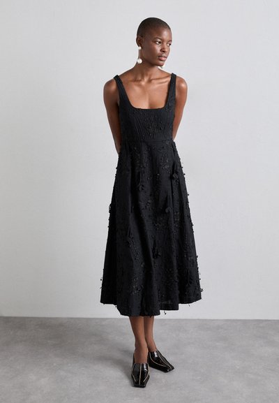 Robe noire sans manches avec un décolleté carré, ornée d'une broderie florale texturée et d'une jupe évasée fluide, associée à des chaussures noires brillantes.