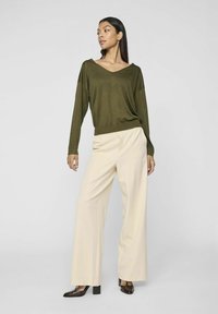 Olive green, długi sweter z dekoltem w serek i długim rękawem w połączeniu z jasnymi spodniami o szerokich nogawkach oraz ciemnobrązowymi szpilkami, tworząc kontrastowy wygląd.