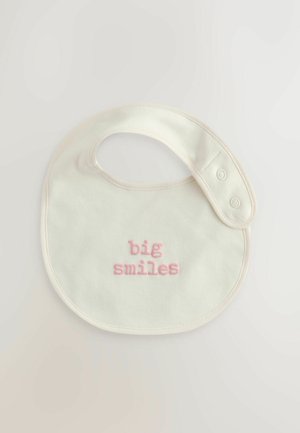 Pamučni podbradnik u boji slonovače zaobljenog oblika, sa vezom ružičastog teksta koji glasi "velike osmijehe". Dva dugmeta za kopčanje sa strane.