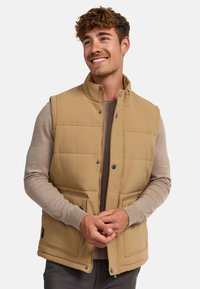 Gilet rembourré beige avec un col haut, des boutons-pression et deux poches avant. Porté sur un pull marron clair, présenté sur un fond neutre.