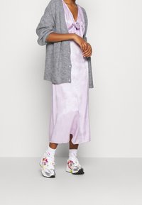 Person som bär en lila satin slipklänning, en grå stickad cardigan, vita strumpor och mångfärgade vita New Balance-sneakers, stående mot en enkel vägg.