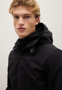 Schwarze Jacke mit Reißverschluss vorne, Kapuze und zwei großen Fronttaschen. Sie verfügt über eine glatte Textur und silberne Hardware-Details.