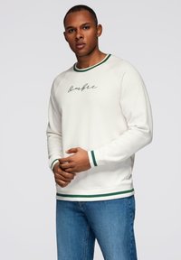 Man som bär en vit sweatshirt med grön kant och "Ombre"-skrift, tillsammans med blå jeans, står mot en enfärgad ljus bakgrund.