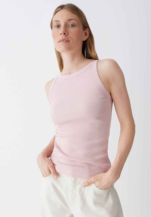 Jonge vrouw met steil haar, gekleed in een roze mouwloos geribbeld topje en witte broek, staand met handen in de zakken tegen een witte achtergrond.