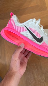 Mână ținând o sneaker Nike ZoomX alb și roz strălucitor cu un swoosh negru, pe un fundal din podea din lemn.