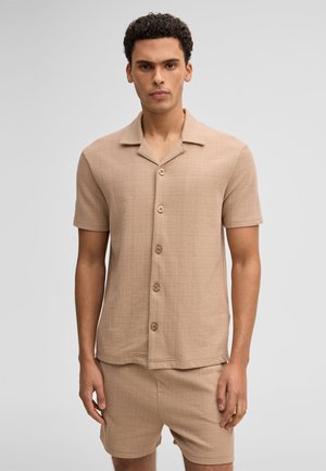 Młody mężczyzna o kręconych włosach, ubrany w beżową, teksturowaną koszulę z krótkim rękawem zapinaną na guziki oraz dopasowane szorty, stojący na jednolitym tle.