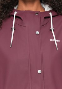 Casaco impermeável cor vinho com capuz, detalhes em prata, cordões ajustáveis e texto da marca. Textura suave com um ligeiro brilho.