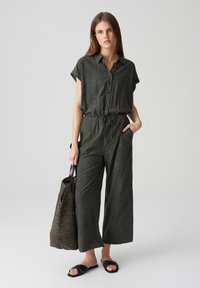 Dunkelgrüner Jumpsuit mit kurzen Ärmeln, Knopfleiste, tailliertem Schnitt und weit geschnittenen Hosenbeinen. Kombiniert mit einer schwarzen gewebten Tragetasche und schwarzen Sandalen.