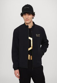 Svart zip-up jacka med guld EA7-logotyp på bröstet. Bärs över en svart och guldmönstrad T-shirt. Svart keps med vit text.