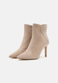 Bottines en suède beige avec des orteils pointus et des talons fins, présentant une texture lisse et un design de couture latérale courbée.