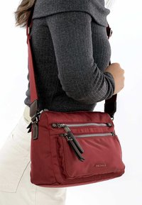 Červená látková crossbody taška s nastavitelným popruhem, vybavená stříbrnými zipy, dvojicí kapsiček na zip vpředu a decentním logem značky po straně.
