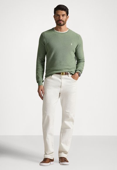 Green knit sweater with a crew neck, paired with light cream jeans. Brown shoes and a belt complement the outfit. Minimalist logo detail visible.