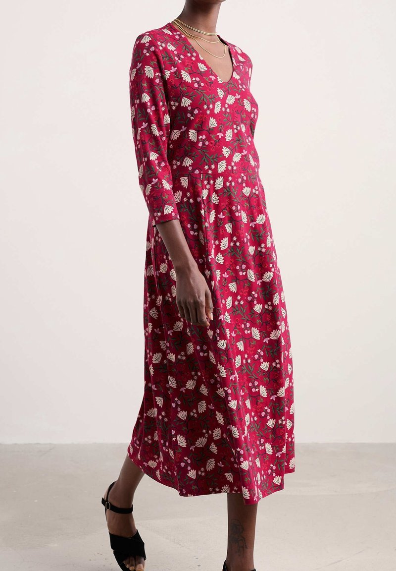 Femme portant une robe midi rouge avec des motifs floraux blancs et des manches trois-quarts, associée à des sandales noires ouvertes à l'avant et des colliers en or délicats.