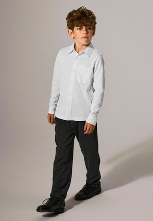 Camicia bianca a maniche lunghe con taschino sul petto, abbinata a pantaloni grigi scuri e scarpe nere, indossata da un bambino con capelli ricci.