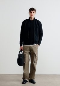 Νεαρός άνδρας όρθιος, φοράει μαύρο μπουφάν με φερμουάρ, μπεζ παντελόνι cargo, μαύρα παπούτσια, κρατώντας μια μαύρη τσάντα χειρός μπροστά σε απλό λευκό φόντο.