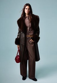 Hnědý kožený kabát s kožešinovým límcem, odpovídající hnědá croptopová bunda se zapínáním na knoflíky, široké hnědé kalhoty a malá červená kabelka.