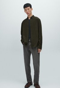 Camicia verde oliva con bottoni, dal tessuto morbido, indossata sopra una maglia scura, abbinata a jeans grigi e scarpe marroni. Design semplice con accenti minimi.