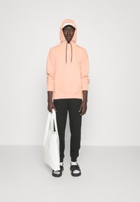 Světle růžová mikina s tkaničkami, černé tepláky, bílá taška a černé sandály nošené s bílými ponožkami; model stojí na jednobarevném pozadí.