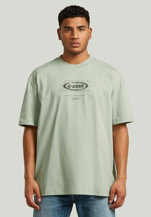Młody mężczyzna z krótkimi kręconymi włosami, ubrany w jasnozielony, oversize’owy t-shirt z szarym napisem oraz casualowe niebieskie dżinsy na jednolitym tle.