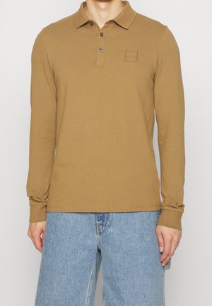 Mann trägt ein braunes langärmliges Poloshirt mit Knopfleiste und hellblaue Jeans, steht vor einem schlichten Hintergrund.