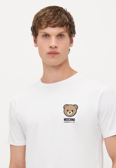 Jaunas vyras vilki baltą marškinėlį, kuriame kairėje krūtinės pusėje yra mažas meškiuko piešinys ir užrašas "Moschino Underbear".