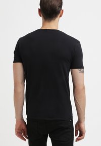 Mand iført en ensfarvet sort T-shirt med korte ærmer og sorte bukser, der står med ryggen til kameraet mod en hvid baggrund.